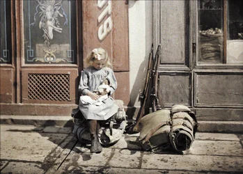 Ein kleines Mädchen spielt mit ihrer Puppe; zwei Gewehre und ein Rucksack liegen neben ihr auf dem Boden, Reims, Marne, Frankreich, 1917 (Autochrom)